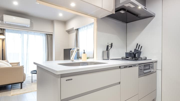 Modern kitchen view with a white countertop island featuring a sink and faucet, a gas cooktop, and a glimpse of the living room (sofa and windows) in the background.