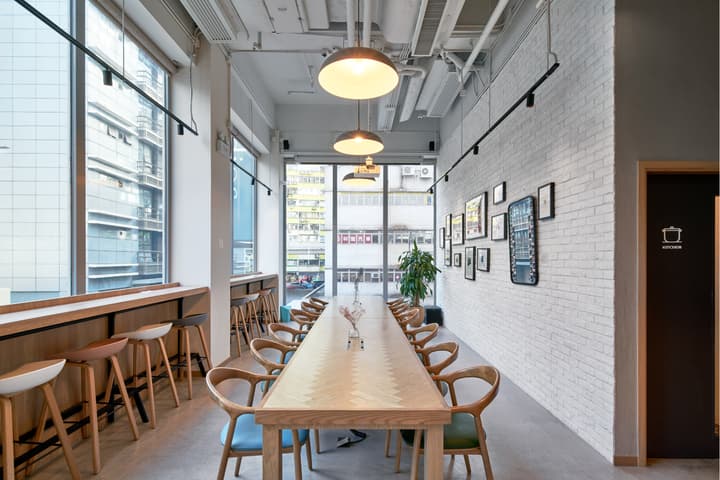 A well-lit communal dining area with a long wooden table featuring a herringbone pattern, matching chairs, bar stool seating along a window, and gallery frames on a white brick wall.