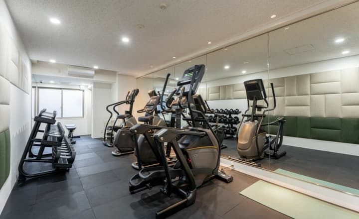 Shared fitness center area with a mirrored wall, black rubber floor, featuring various cardio machines and a rack of dumbbells.