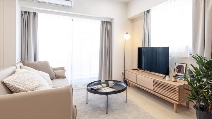 Furnished living room with a neutral-toned sofa, a modern wooden TV stand, a flat-screen television, a round black coffee table, and large windows with sheer and grey curtains.