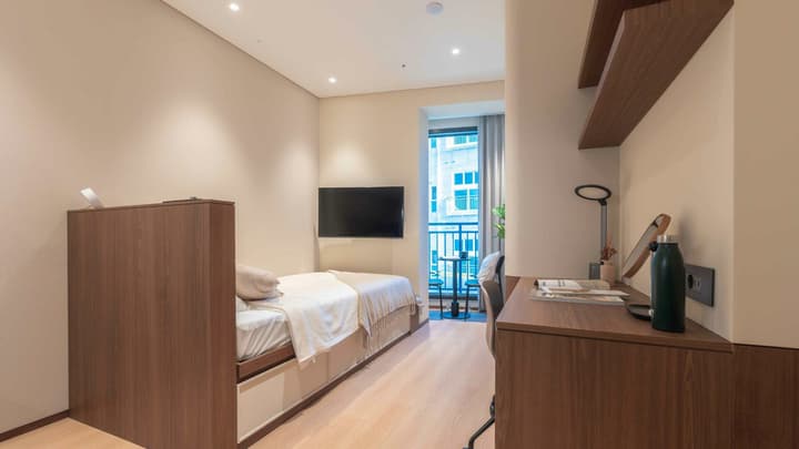View of a compact, well-lit studio apartment from the perspective of the entryway, showing the single bed with under-bed storage, a wall-mounted TV, and a functional built-in wooden desk with shelving.