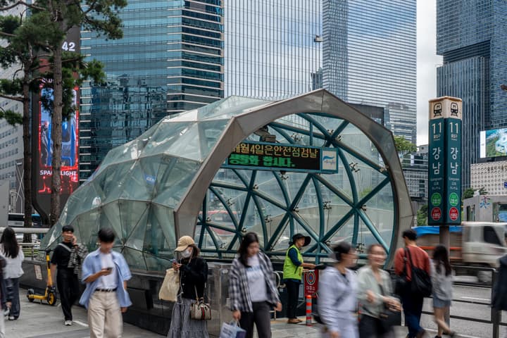 A modern, geodesic glass dome structure serving as an entrance/exit for the Gangnam Subway Station in Seoul, with blurred people walking on the sidewalk around it.