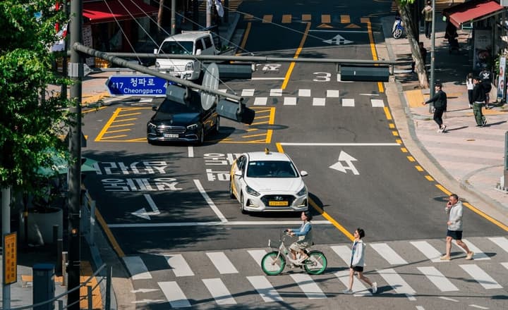 Overhead view of a busy street intersection (Cheongpa-ro, Seoul) with traffic, including a white taxi, pedestrians walking on a crosswalk, and a person riding a bicycle.