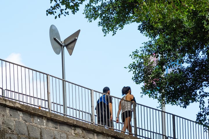 A man and woman in athletic wear walking along a raised walkway with a metal railing, stone retaining wall, and lush green tree canopy overhead.