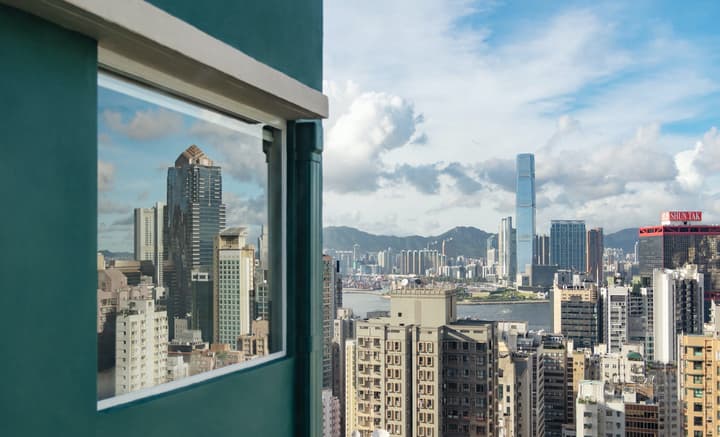 High-angle view of the Hong Kong skyline and Victoria Harbour, framed by a teal exterior wall and a window reflecting the towering buildings and clouds.