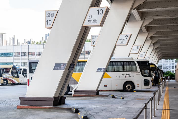 A loading bay at a Korean intercity bus terminal with white structural pillars displaying route signs (e.g., Gumi, Sangju, Pohang) and a line of parked express buses.
