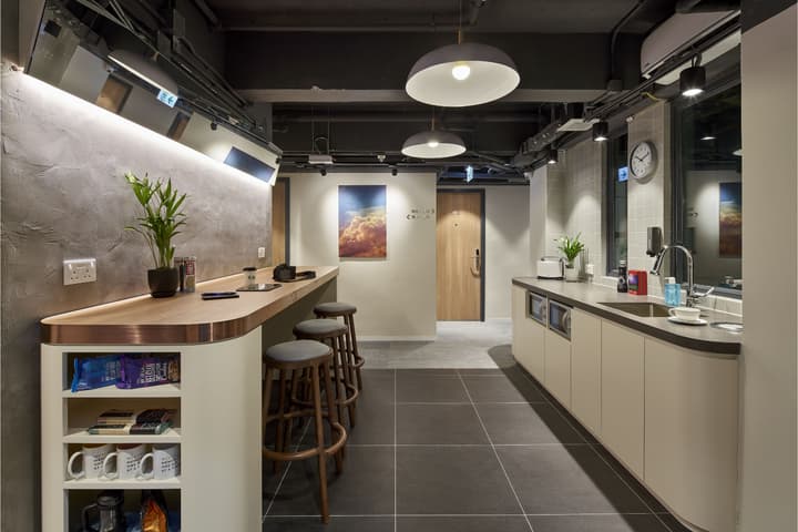 A modern communal pantry with a dark color palette, featuring a long wooden breakfast bar with three stools and a kitchenette area with a sink and built-in microwaves.