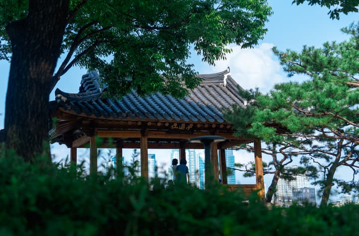 A traditional Korean wooden pavilion (jeongja) with a tiled roof, framed by lush green trees and foliage, with a modern city skyline visible in the hazy distance.