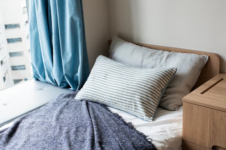A close-up shot of a cozy bed with light blue and striped pillows, a dark blue throw blanket, next to a wooden nightstand and a window with blue curtains.