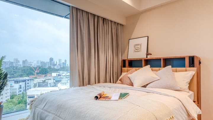 Premium suite bedroom showing a queen bed with a tan upholstered headboard, integrated shelving, and a large window with city and tree views.