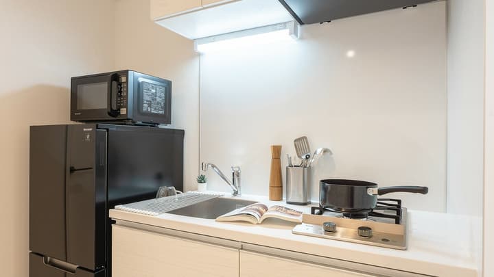Compact kitchen area with a small single-burner cooktop, white countertop, sink, a black mini-fridge, and a black microwave, with cooking accessories and a cookbook on the counter.