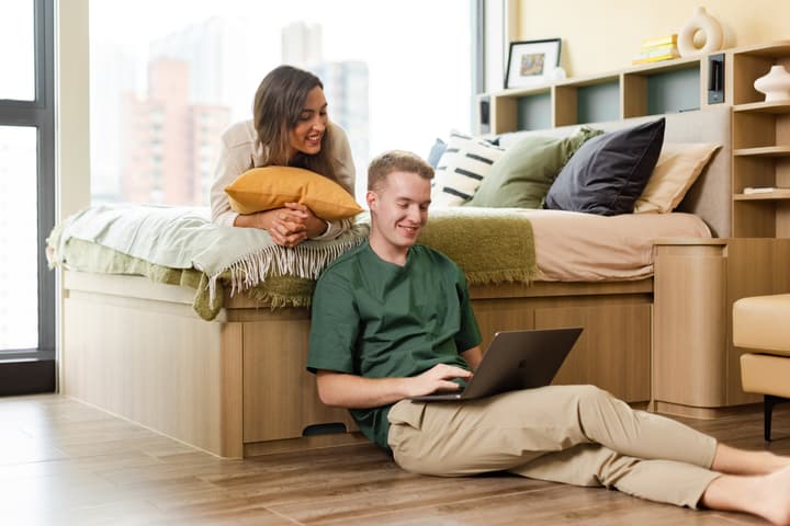 A happy couple relaxing in a modern studio apartment; the man is sitting on the wood floor using a laptop, and the woman is leaning over the storage bed with a window view behind them.
