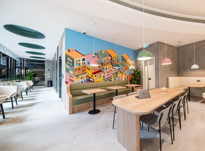 A vibrant co-working or communal lounge area with a long shared table, green booth seating, and a striking colorful mural depicting houses on a hill.