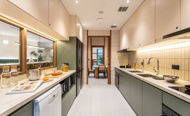 Sleek communal kitchen with parallel counters, sage green lower cabinets, a double sink, black integrated fridge, and view into the adjacent dining area.