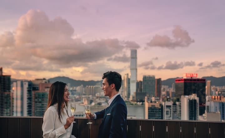 A well-dressed couple toasting with champagne flutes on a rooftop at sunset, with a vibrant sky over the Hong Kong city skyline in the background.