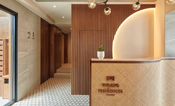 Stylish and modern apartment lobby with a curved, textured reception desk featuring the 'Weave Residences' logo, wood slat accent walls, and warm lighting.