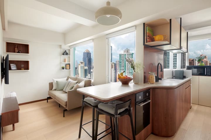 Bright, modern studio apartment with an open-plan kitchenette, small sofa living area, and large windows offering a city skyline view.
