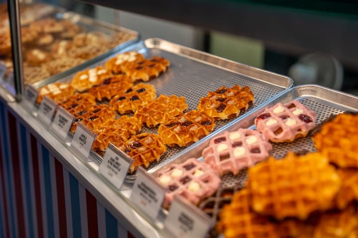 A close-up display of different flavors of freshly made croffles (croissant-waffles) on metal trays, including glazed, raspberry, and plain varieties with price tags.