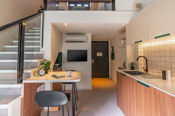 Lower level of a loft suite showcasing a compact kitchenette with white and wood-look cabinets, a dining counter for two, and a modern staircase leading to the upper floor.