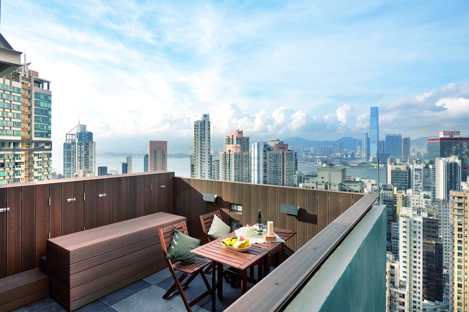 Stunning rooftop terrace with a wooden dining set and built-in bench, offering a panoramic view of the Hong Kong city skyline and harbor under a blue sky.