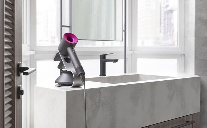 A modern bathroom counter with a large, square concrete sink, black faucet, and a purple Dyson hair dryer on a stand next to a window with a city view.