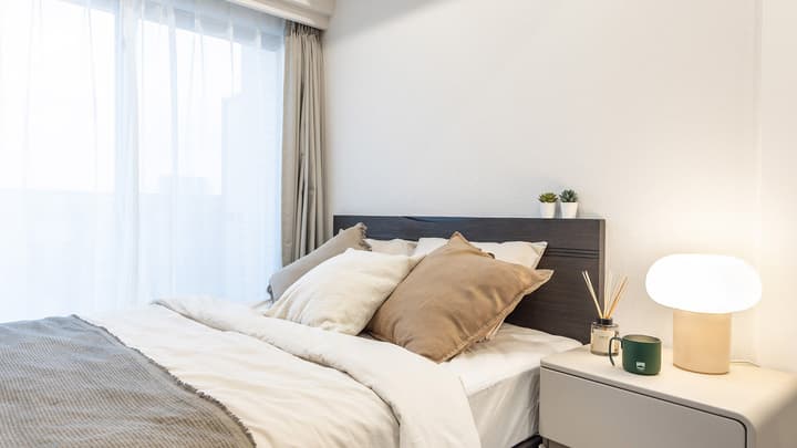 Close-up of a dark wood bed headboard and a light-colored bedside table with a modern white lamp, mug, and reed diffuser, next to a large window with sheer and blackout curtains.