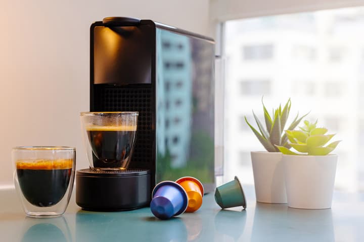 Close-up of a modern espresso machine with coffee capsules and glass cups on a countertop beside small potted plants in a bright apartment kitchen.