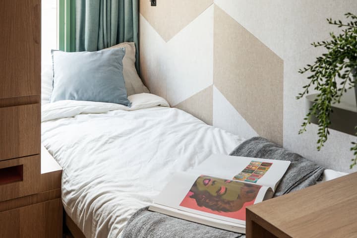 A close-up view of a single bed with white bedding and a light blue velvet pillow. An open art book displaying a colorful portrait rests on a gray throw blanket. The headboard wall is covered in a decorative geometric pattern of light beige and white.