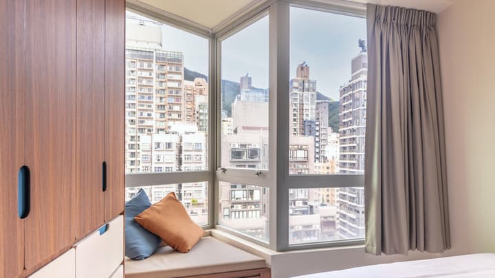 Interior view of a modern studio unit with a large corner window, built-in window seat/bench, and a tall, wood-grain storage cabinet on the left, offering a dense city view.