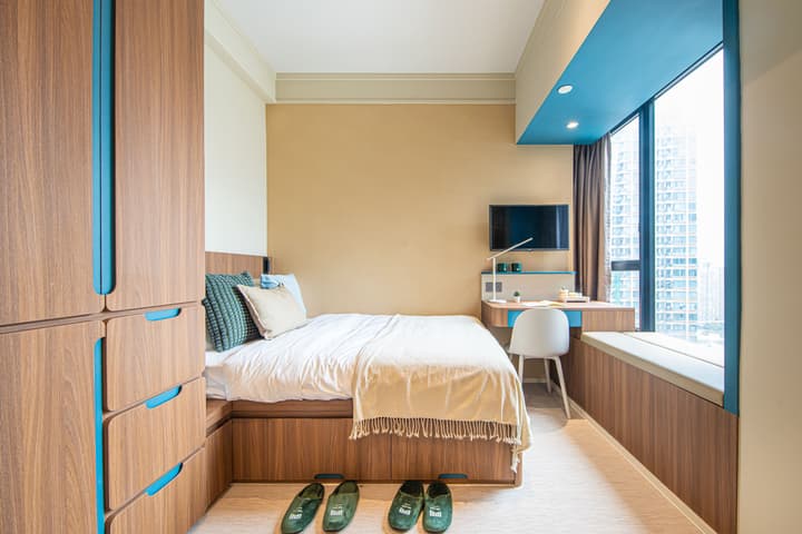 A view of a compact bedroom with warm beige walls and built-in wood-grain furniture accented with light blue. The room features a single bed, a large integrated storage cabinet with drawers, a small desk area with a wall-mounted TV, and a window seat with a city view.
