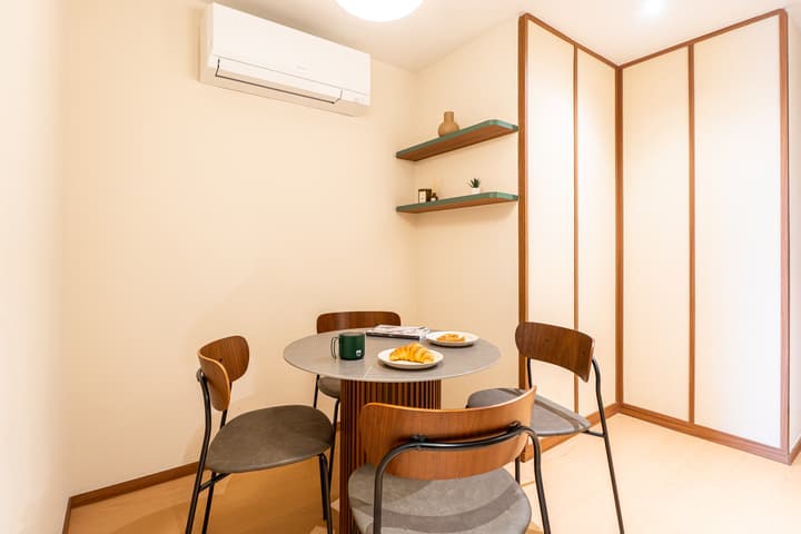 Dining area with a round table on a slatted wood base, four wood-framed chairs, and a wall of neutral wood-framed paneling.
