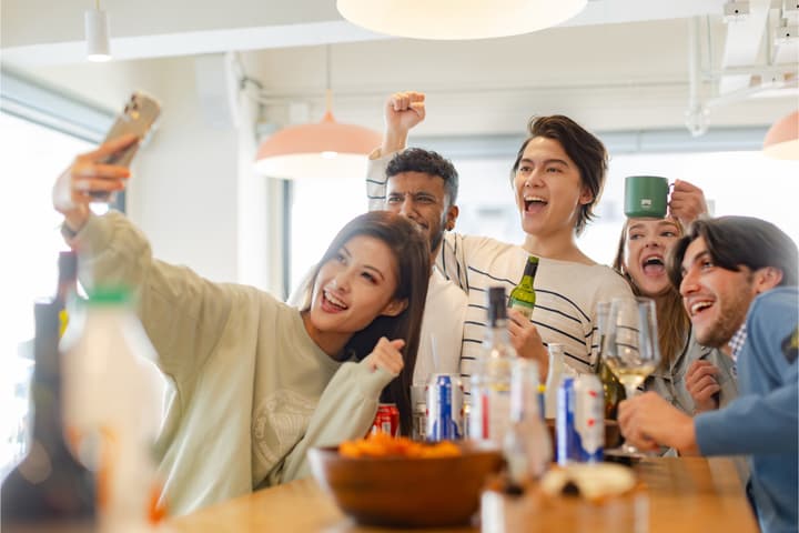 A group of diverse friends celebrating and cheering while taking a festive selfie together around a table filled with drinks and snacks at a party or social gathering.