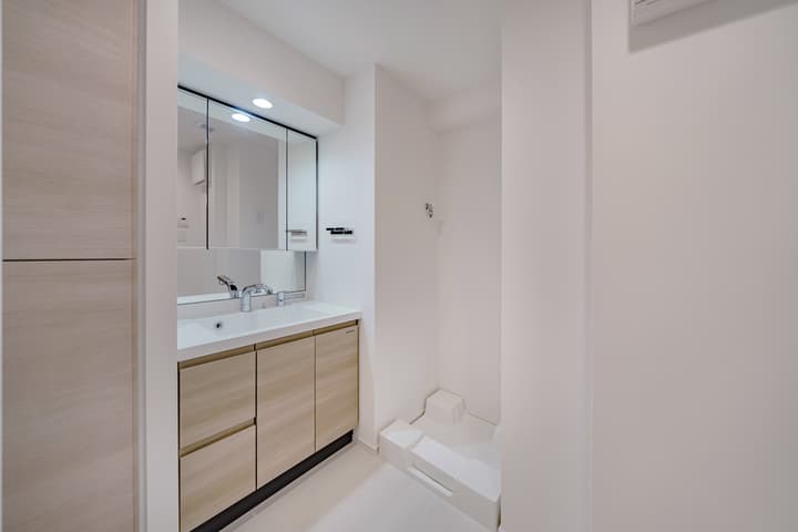 Bathroom vanity with a light wood cabinet, white basin, mirrored medicine cabinet, and a dedicated, tiled floor space to the right for a washing machine hookup.