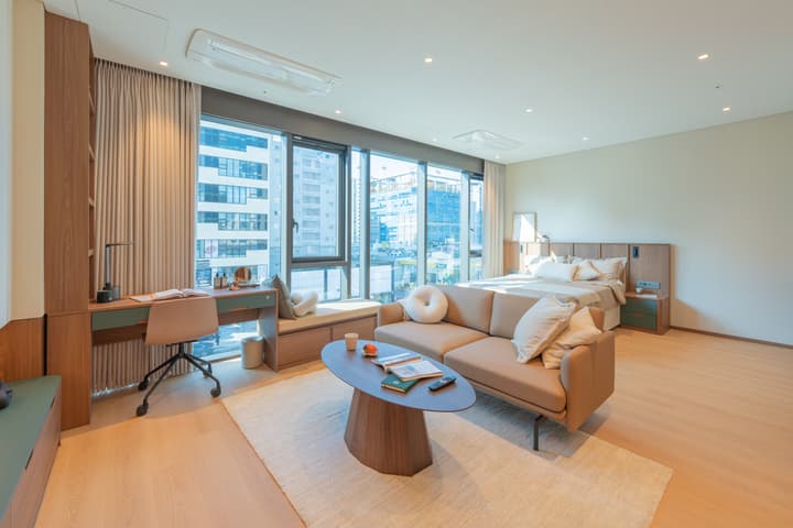 Very wide, bright interior shot of the Junior Luxe Suite, showcasing the generous floor space, queen bed, tan leather sofa, and an integrated desk area next to the expansive floor-to-ceiling windows.