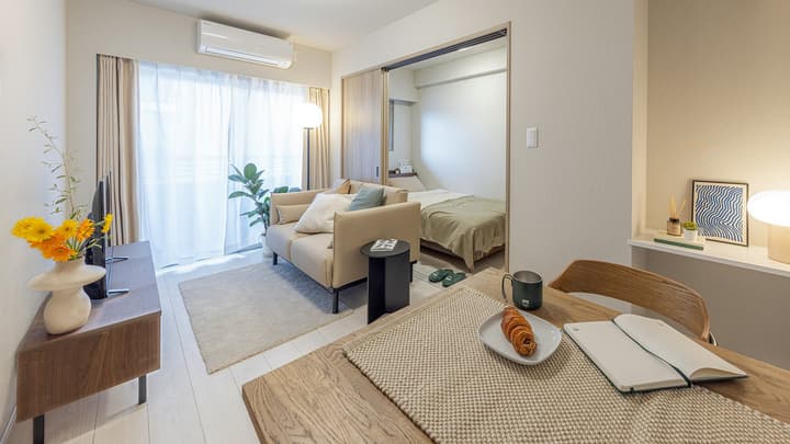 Open-plan living and dining area featuring light wood-style flooring, a light beige sofa, a dining table set for two, and a view into the adjacent bedroom through a sliding partition in Monzennakacho.