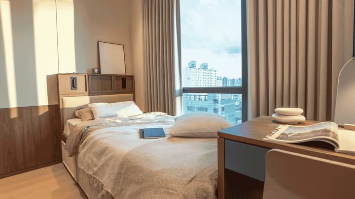 Sunlit studio bedroom with a queen-size bed, a dark wood headboard with built-in storage, and a minimalist writing desk next to a large window overlooking a city view.