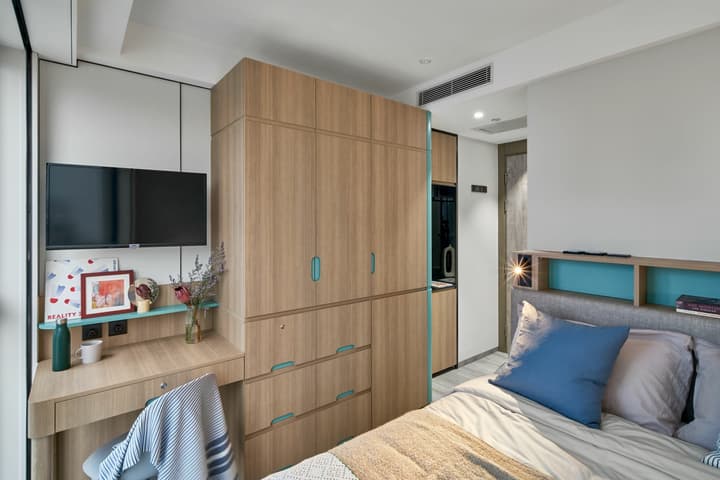 Bright, modern studio apartment interior featuring a queen bed with a turquoise shelf headboard, a wall of wood-grain built-in cabinetry including a wardrobe and fridge, a desk area with a mounted TV, and light wood furnishings.