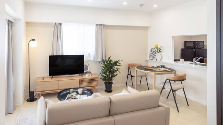 Furnished living room with a neutral-toned sofa, light wood TV stand, and a round black dining table with two modern chairs, with a pass-through opening to the kitchen.