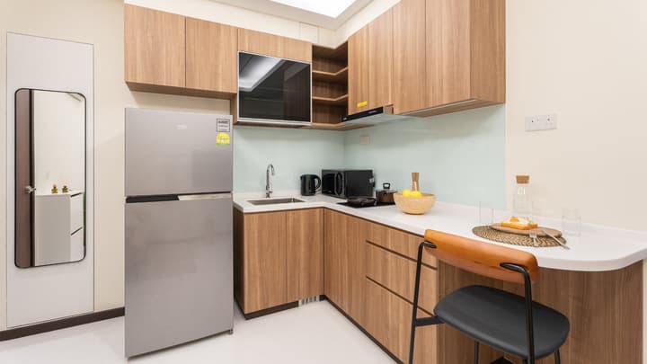 L-shaped apartment kitchen with light wood-look cabinets, a stainless steel refrigerator, white countertops, and a clear glass backsplash.