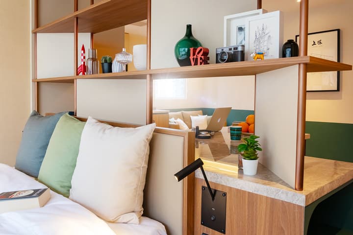 lose-up view of a modern bedroom's built-in desk area, featuring a marble-look countertop, a laptop, and a wooden shelf divider above decorated with a red rocket, a vintage camera, framed art, and various decorative objects.
