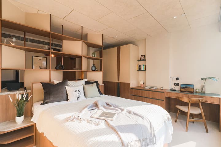 Bright, modern one-bedroom suite interior featuring a queen bed with a light throw blanket, a built-in wooden shelf divider behind the bed, a wardrobe, and a separate desk and chair with ample natural light.