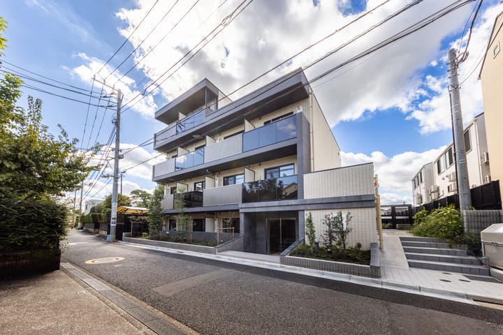 Exterior street-level view of a modern, three-story apartment building with dark grey and light beige accents, glass balcony railings, and simple landscaping under a blue, cloudy sky.