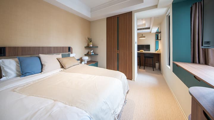 View from a queen-sized bed looking into the adjoining living area of a studio apartment. The bedroom has wood and beige tones, while the living area features a bar-height table and stools, a TV, and shelves in a darker, contrasting color scheme.