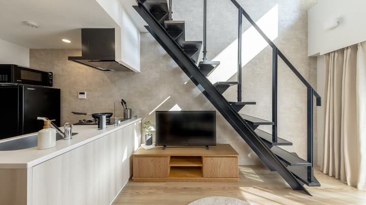 View of the living and dining area featuring a black open-tread steel staircase leading to the loft, a modern kitchen counter on the left, and a TV on a wooden cabinet.