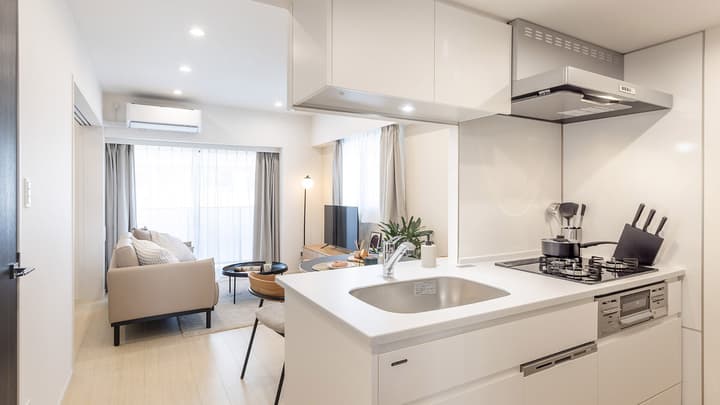 Modern, bright white kitchen with recessed lighting, a built-in cooktop, and a stainless steel sink, overlooking the open-concept living and dining area in the background.