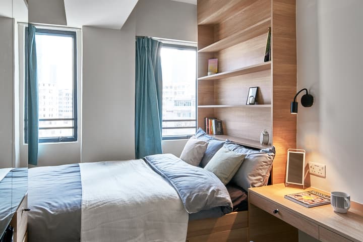 A modern studio apartment bedroom featuring a queen-sized bed with blue and white bedding, built-in wooden shelving, a small integrated desk, and two tall windows with blue curtains.