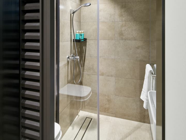 Close-up view of a modern walk-in shower stall with a clear glass door, featuring light beige, textured stone-look wall tiles, a chrome shower head, and a vertical linear drain.