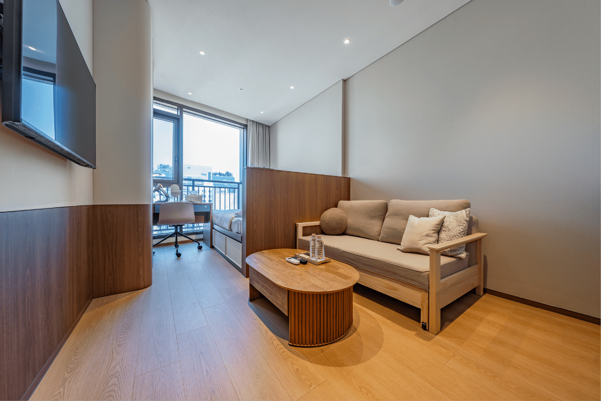 Wide view of a modern studio apartment with a queen bed and a separated sofa bed lounge area featuring a unique adjustable height coffee table, set near a large window.