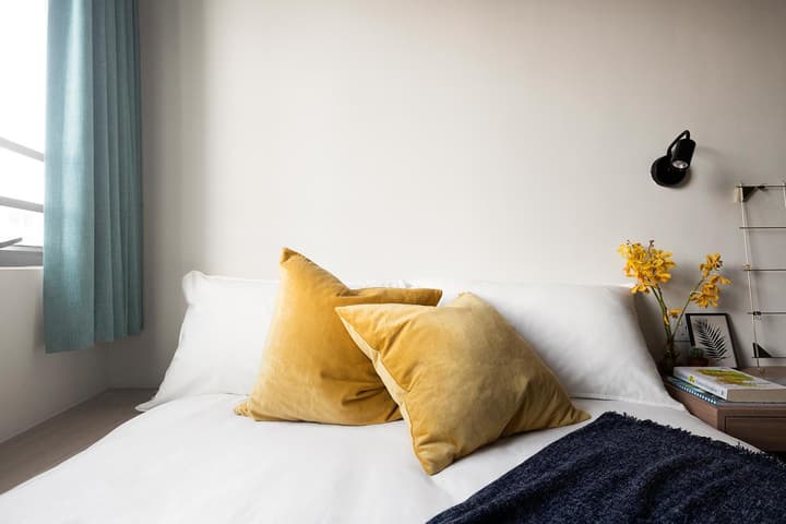 A close-up of a double bed in a studio apartment with crisp white bedding, two prominent mustard yellow velvet pillows, a navy blue throw, and yellow flowers on a wooden nightstand.