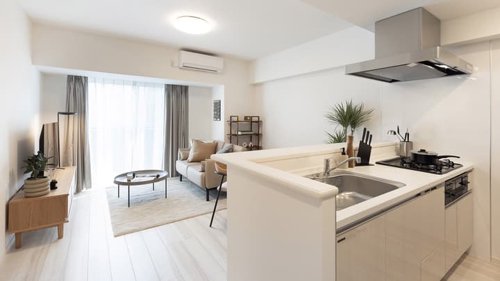 Wide-angle view of a bright, open-concept living room and kitchen, showing a light leather sofa, wood TV console, and kitchen counter with a sink and stovetop.